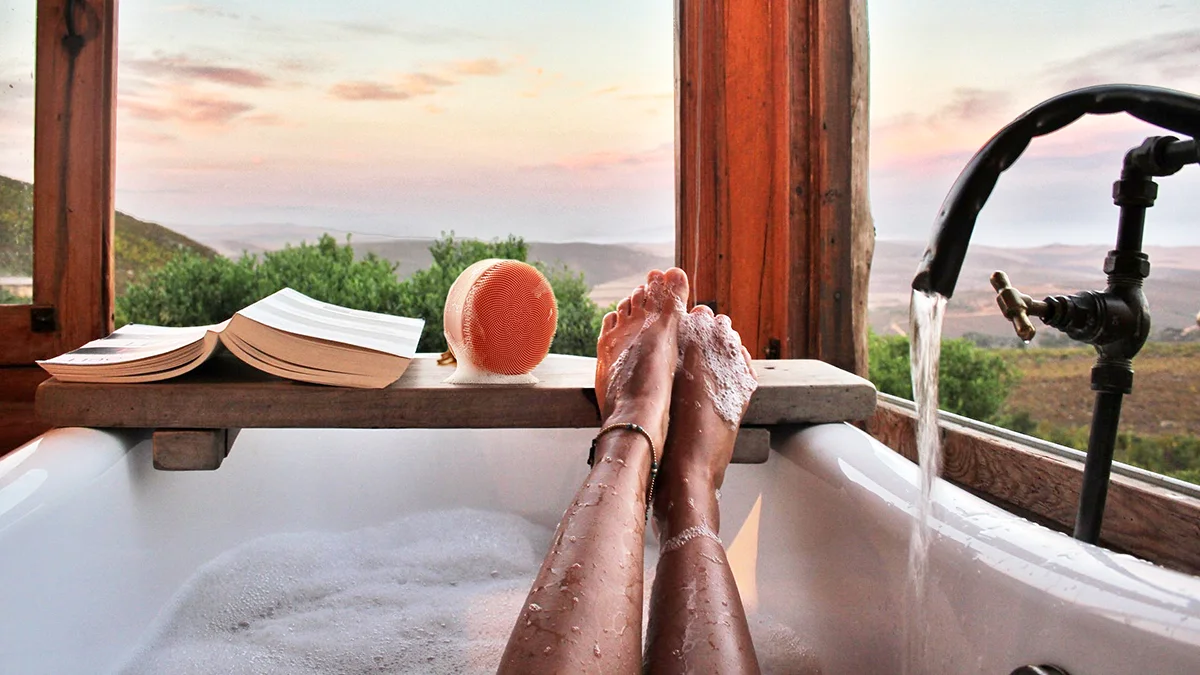 A woman in a bubble bath with her feet up, overlooking a gorgeous sunset in a glass bathroom, with FOREO LUNA 4 body ready to use